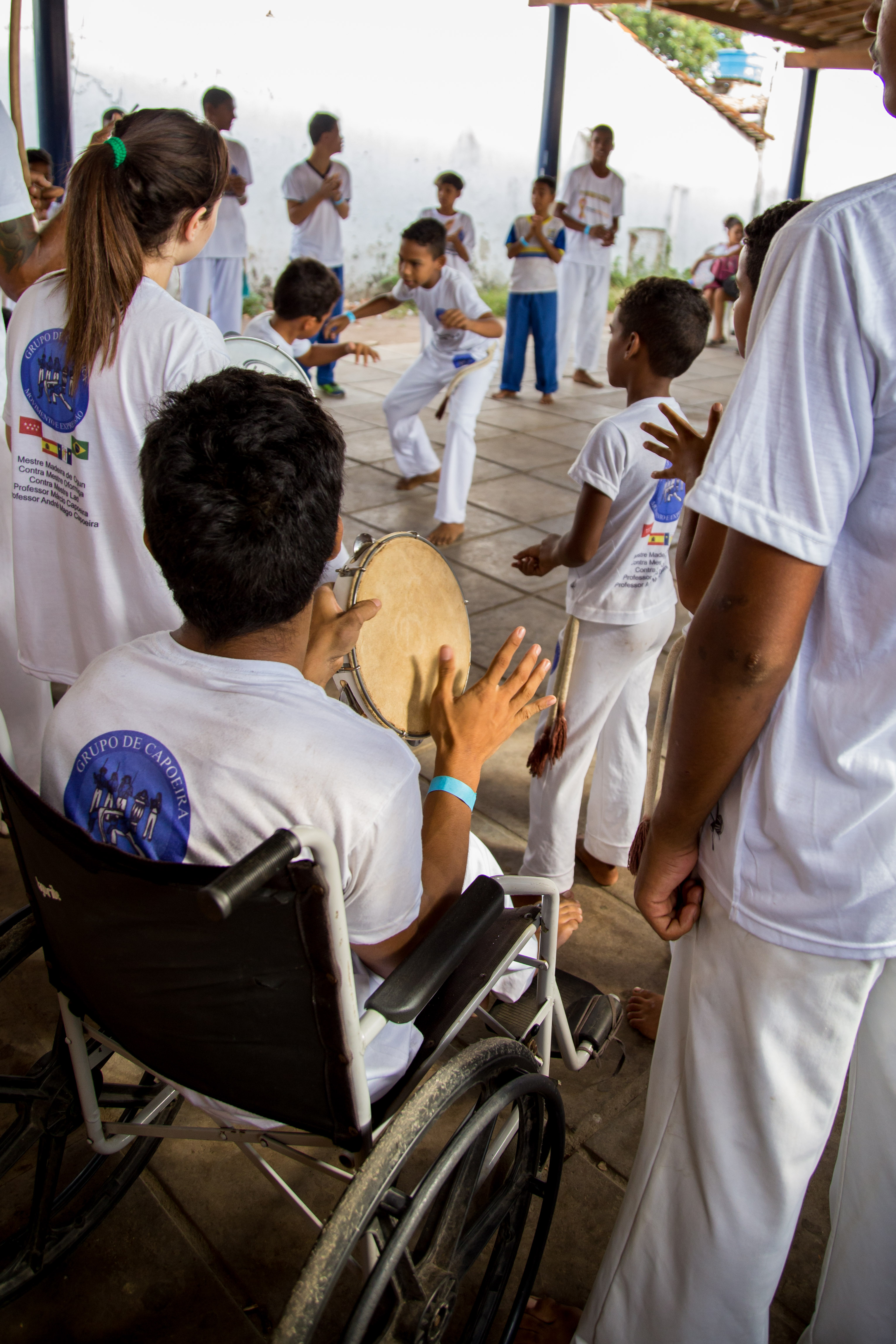 Um estudante com deficiência física está sentado em uma cadeira de rodas e é mostrado de perfil tocando um pandeiro. Ao seu redor, alunos jogam capoeira. Todos vestem roupas brancas.