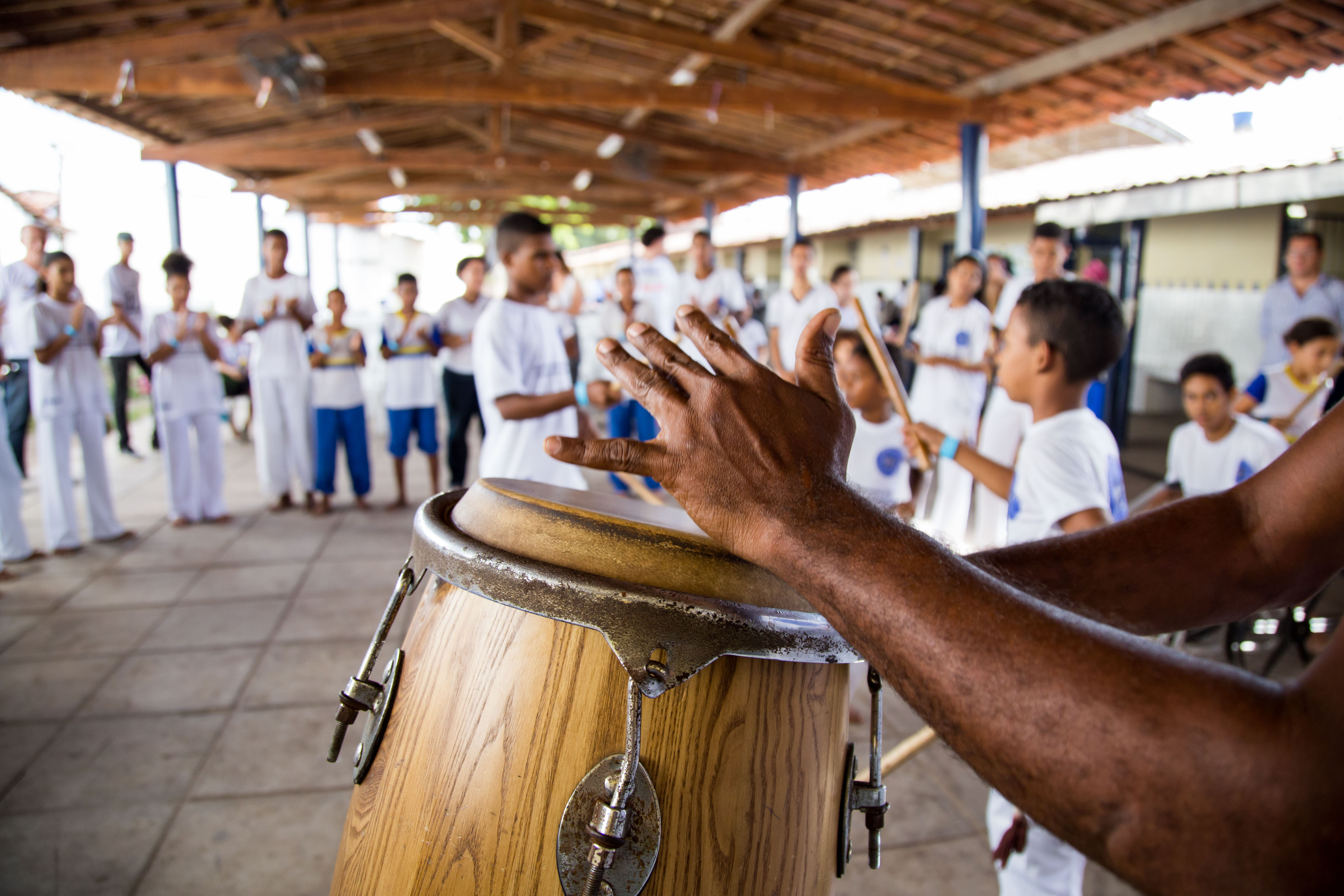 Mão toca o instrumento atabaque (tambor alto), enquanto crianças jogam capoeira ao fundo.