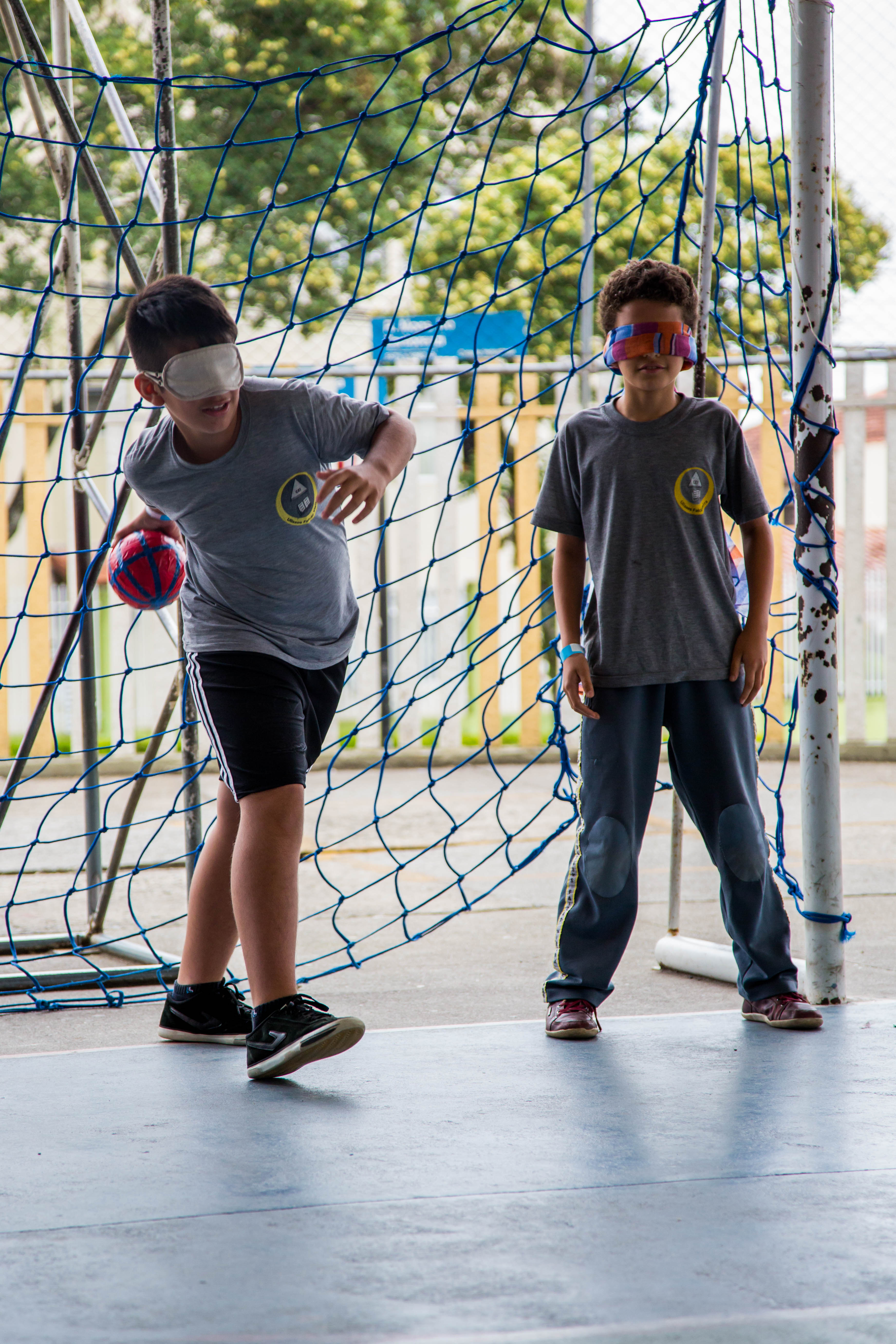 Dois meninos vestem uniformes escolares e têm os olhos vendados. Eles estão à frente de uma rede de gol em uma quadra esportiva. O garoto à esquerda segura uma bola de tamanho médio e faz movimento com o braço para arremessá-la.
