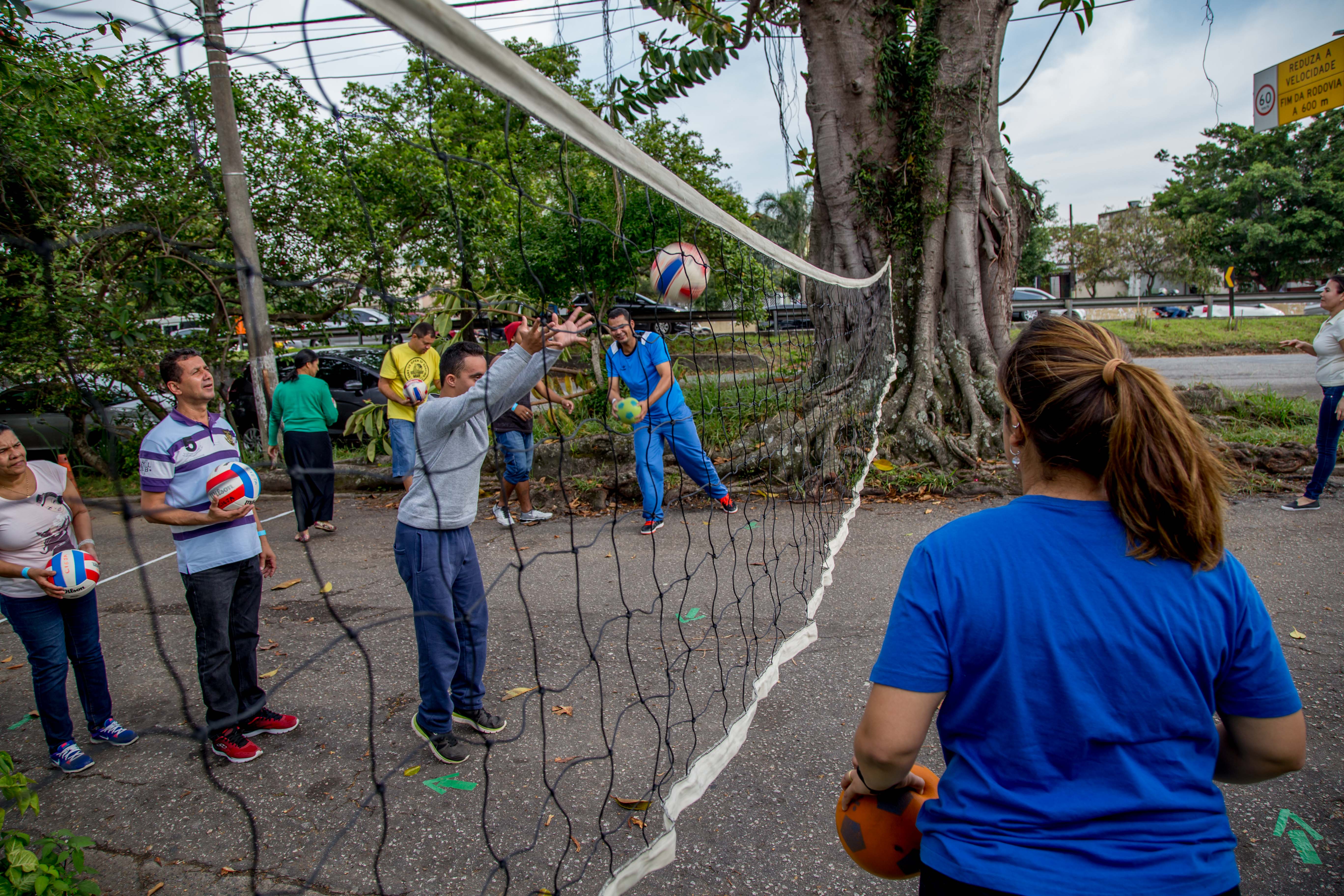 Estudante com deficiência lança bola acima de rede de vôlei pendurada a uma altura baixa na rua. Outros educandos e professores acompanham a atividade ao redor.
