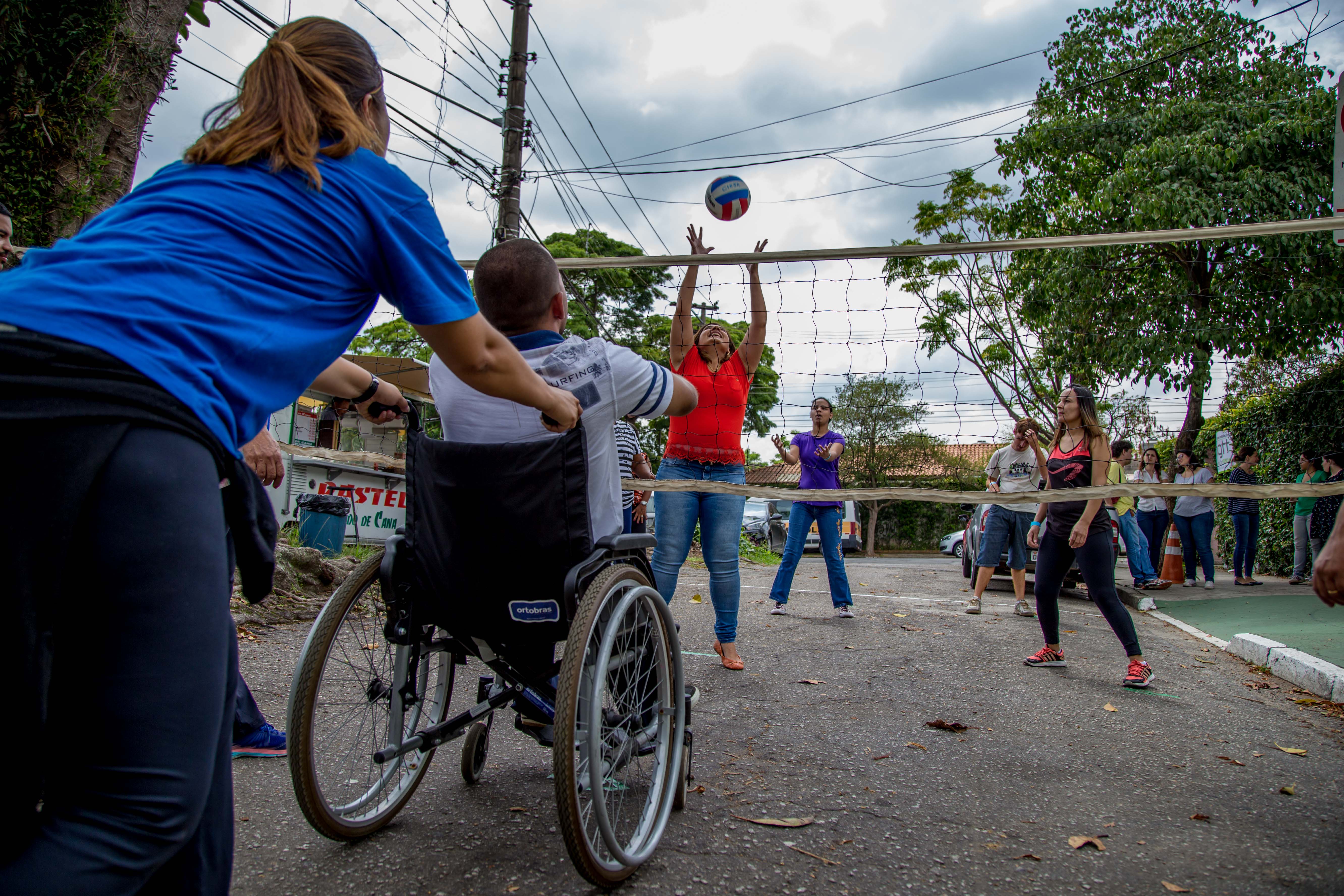 De costas para a imagem, professora empurra cadeira de rodas de aluno com deficiência física que espera recebimento de bola arremessada por outra estudante do lado oposto da rede pendurada na rua.