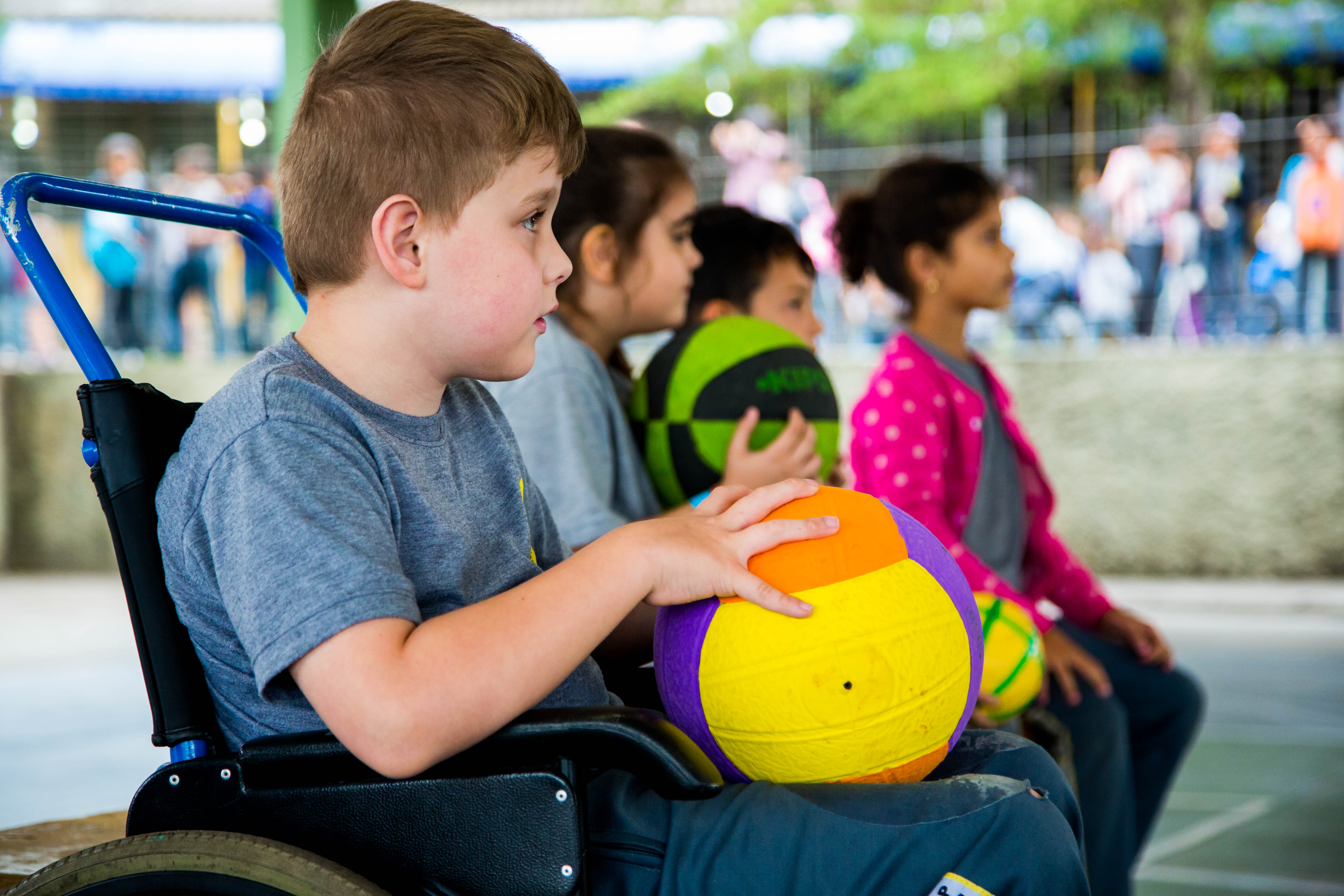 Descrições imagens: Em uma quadra esportiva de escola, um garoto de nove anos em uma cadeira de rodas segura uma bola de vôlei. Ao fundo, outros estudantes estão sentados em cadeiras