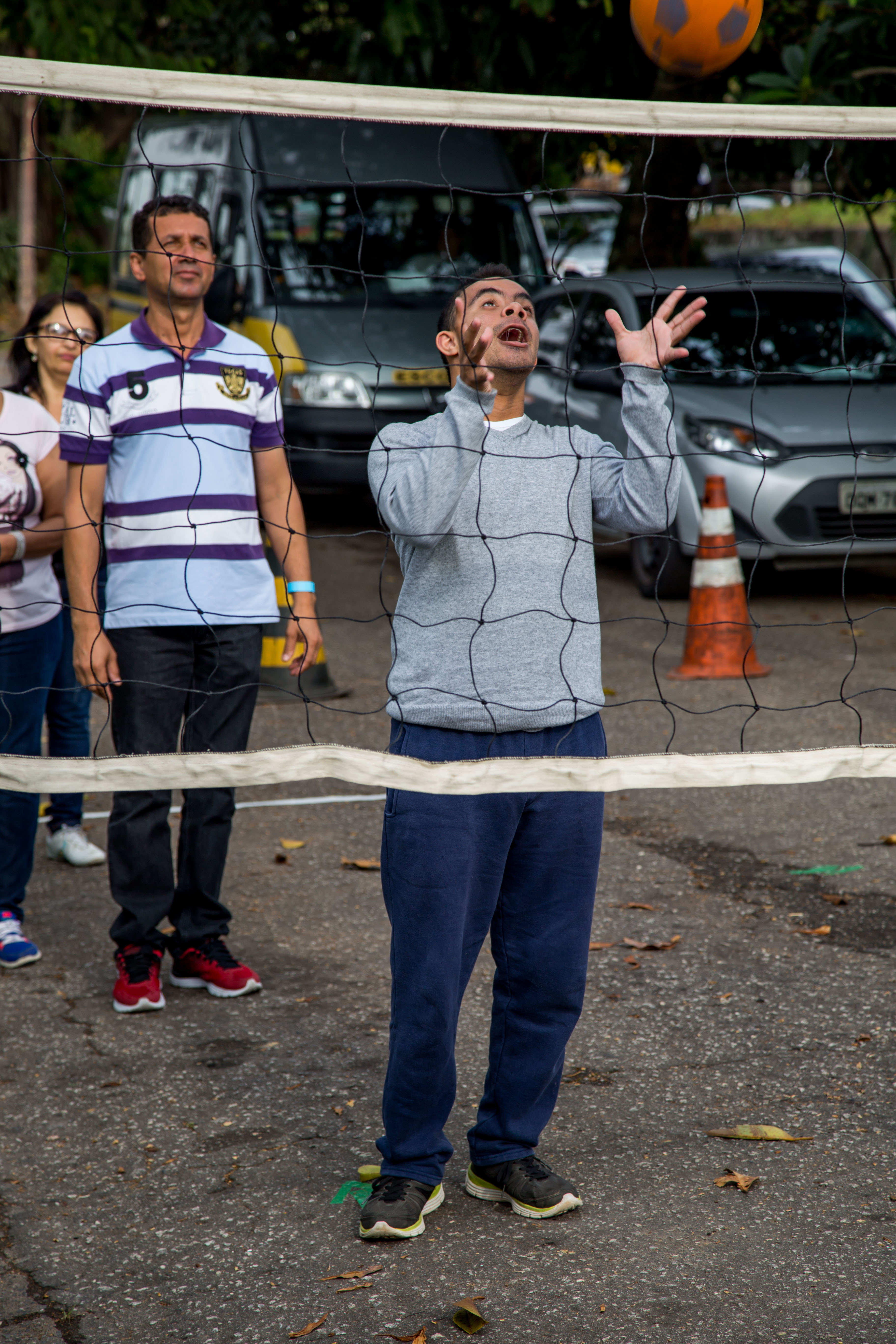 Estudante com deficiência lança bola para cima de rede de vôlei pendurada a uma altura baixa na rua. Ao redor, outros educandos e professores acompanham a atividade.