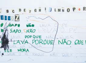 Folha repleta de escritos feitos a mão por uma criança com lápis de cor verde. É possível ler palavras como "Sapo", "não lava porque não" e "mora". Há diversas letras coladas no topo da folha.
