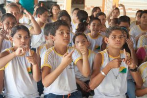 Um grupo de crianças com uniformes escolares faz sinais com as mãos durante aula de Libras