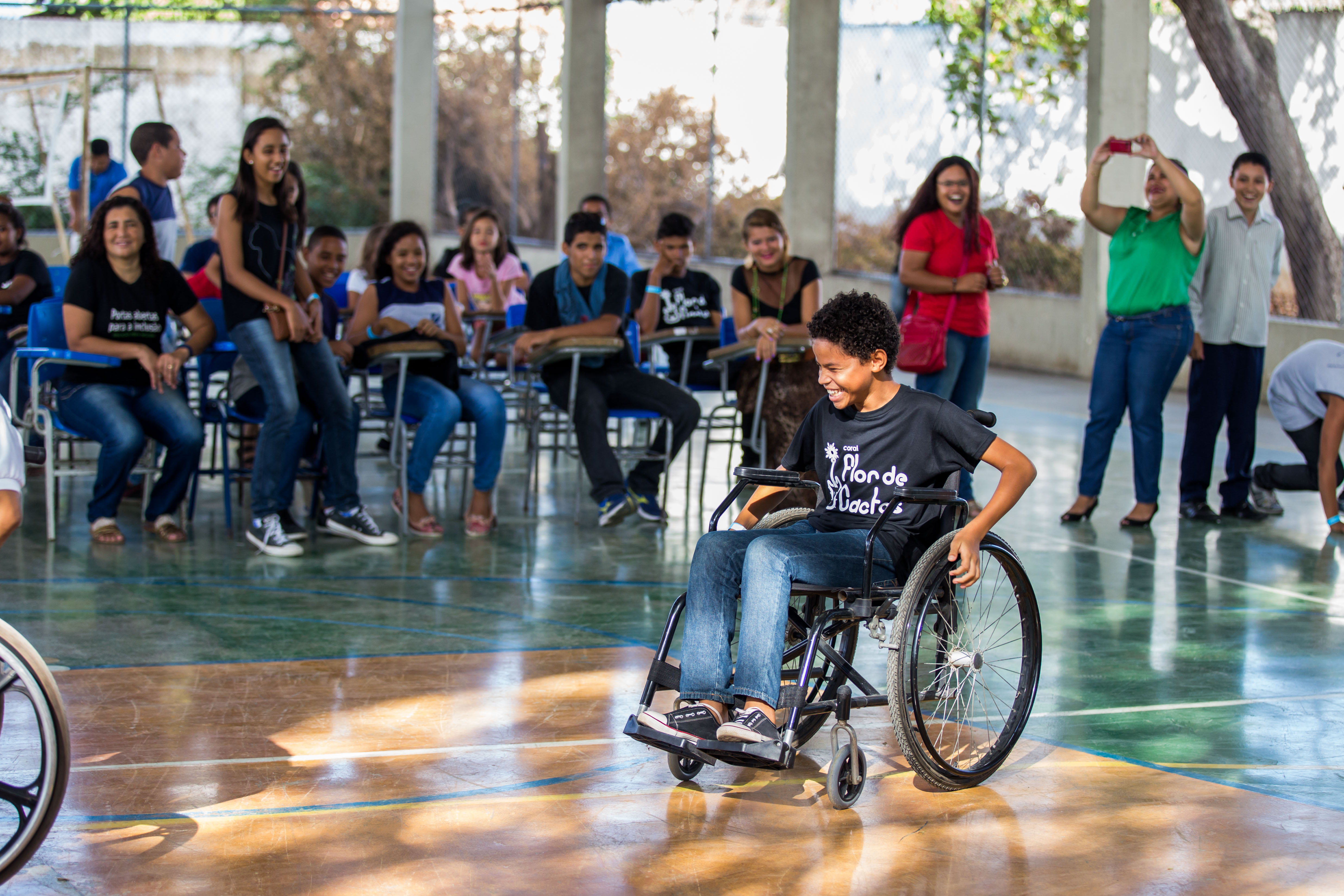 Diversos estudantes entre 10 e 15 anos estão sentados em cadeiras no centro de uma quadra escolar. Um garoto em uma cadeira de rodas se destaca no meio.