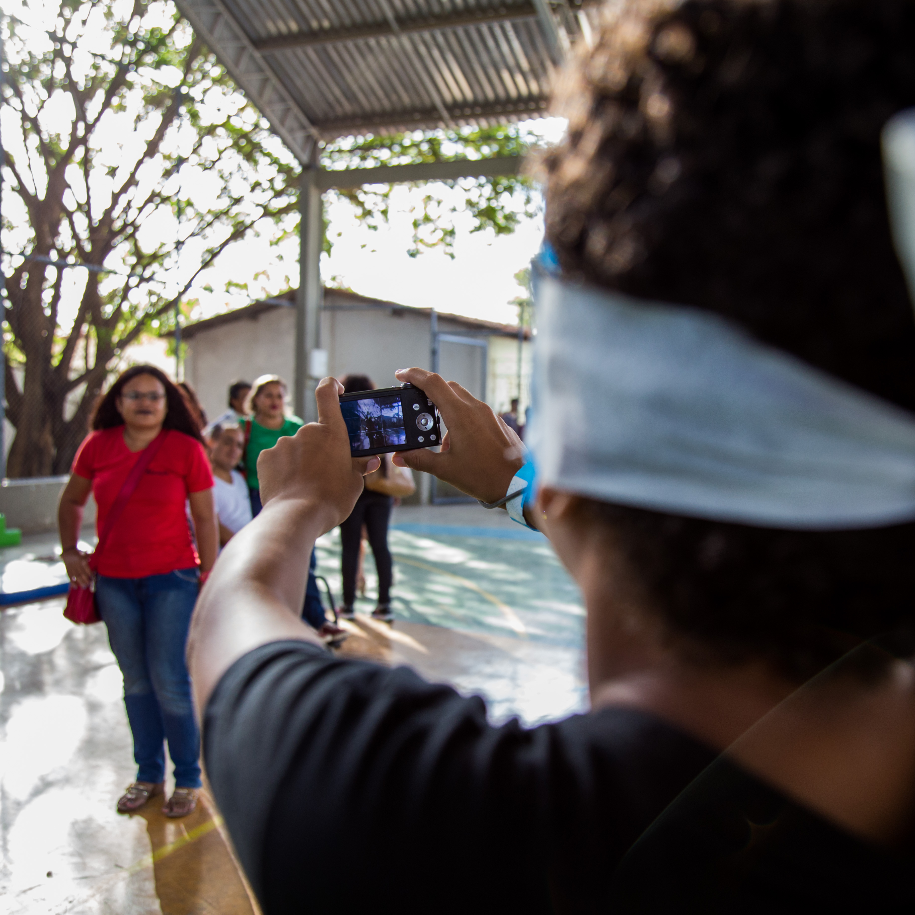 Um aluno vendado segura uma câmera fotográfica digital com as duas mãos. Mesmo sem enxergar, ele procura mirar as pessoas que estão na quadra de esportes.