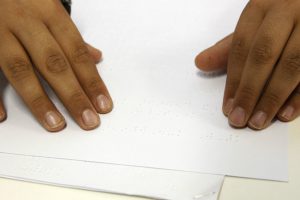 Duas mãos deslizam sobre superfície de folha com impressão braille.