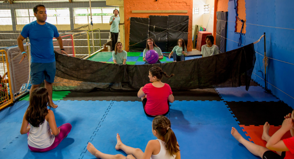 De pé, educador dá instruções para time de jovens em chão acolchoado durante partida de vôlei sentado.