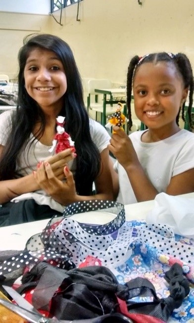 Duas alunas seguram as bonecas que confeccionaram. Elas estão diante de uma mesa repleta de retalhos de tecidos.