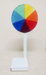 Disco de Newton feito com uma base de madeira, uma haste vertical e um disco fixado na ponta. A circunferência está dividida em sete pedaços. Em cada fatia, os alunos colaram fios das sete cores do espectro da luz.