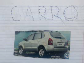 A palavra CARRO escrita por uma criança sobre a imagem de um carro colada em uma página de caderno.