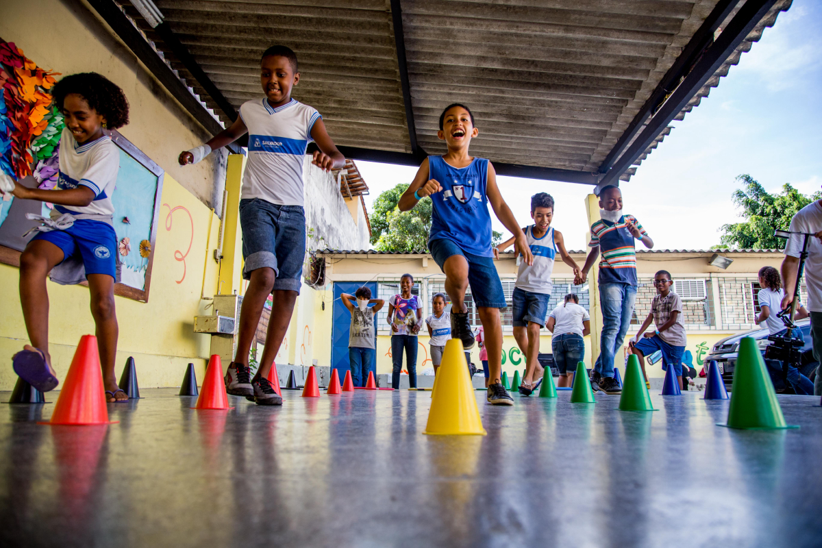 Crianças sorridentes correm entre pequenos cones coloridos dispostos em fila no pátio escolar.