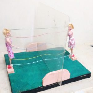 Duas bonecas estão de frente uma para outra. Uma placa de acrílico está fixada verticalmente entre elas, simulando um espelho. Fios de cores diferentes ligam os mesmos pontos nas duas bonecas.