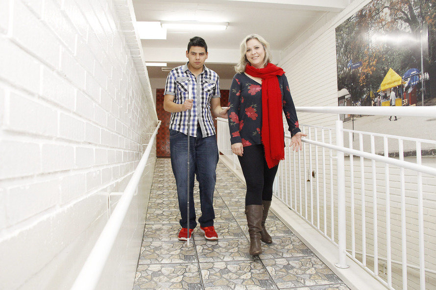 Uma mulher auxilia um estudante cego a se locomover por um corredor. 