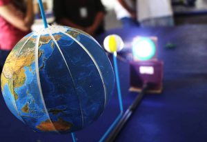Vista do material pedagógico acessível com globo terrestre em primeiro plano e lanterna de led acesa ao fundo.tre e bolas de isopor com as fases da lua.