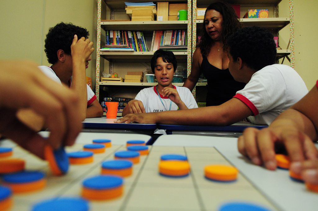 Em sala de recursos multifuncionais, alunos conversam observados por professora. 