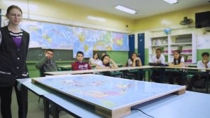 Mapa Mundi interativo fotografado em primeiro plano em sala de aula. Está sobre uma mesa azul. Ao fundo alunos em cadeiras e professora em pé observam o material. Fim da descrição.