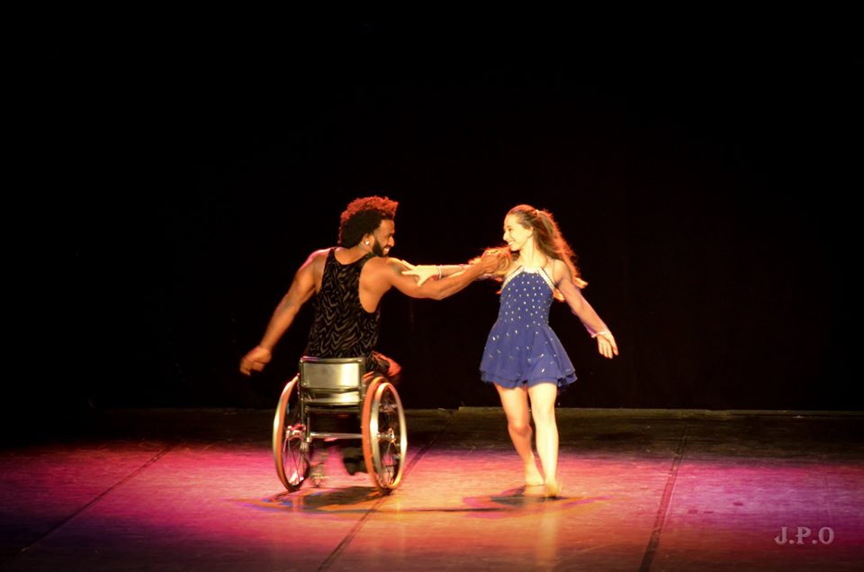 Dançarino sobre cadeira de rodas segura braço de bailarina em pé. Fim da descrição.