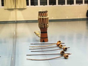 Instrumentos musicais utilizados na capoeira dispostos lado a lado. Os instrumentos são: pandeiros, um atabaque e berimbaus. Fim da descrição.