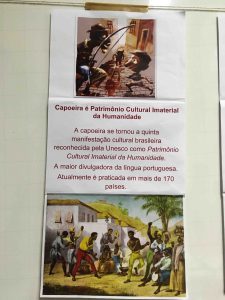 Banner de pesquisa de estudantes sobre a capoeira, com os dizeres "Capoeira é Patrimônio Cultural Imaterial da Humanidade". Fim da descrição.