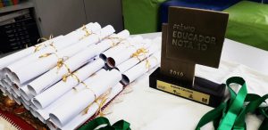 Sobre mesa, certificados enrolados cilindricamente estão agrupados. Ao lado, está o troféu do prêmio, com os dizeres: Prêmio Educador Nota 10 2019 - Arabelle Calciolari. Fim da descrição.