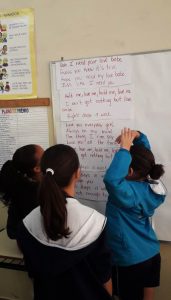 Em sala de aula, três alunas estão de pé em frente a lousa, encarando-a. Nela, está fixada a letra da canção "Eight Days a Week", dos Beatles. Fim da descrição.