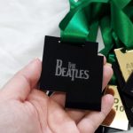Uma mão segura uma medalha de fundo preto com os dizeres: The Beatles. Ao fundo, vemos outras medalhas com o lado dourado para cima, com os dizeres inseridos: Aluno Nota 10 2019. Fim da descrição.