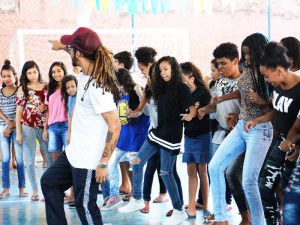 Na quadra escolar, estudantes realizam atividade artística de hip hop. Eles estão reunidos e movimentam a perna da mesma forma, realizando um passo de dança. Fim da descrição.