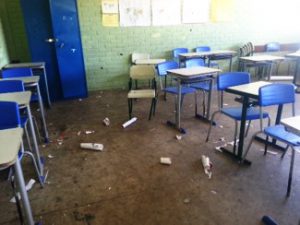 Sala de aula escolar em más condições, com as carteiras bagunçadas, um armário quebrado e lixo no chão. Fim da descrição.