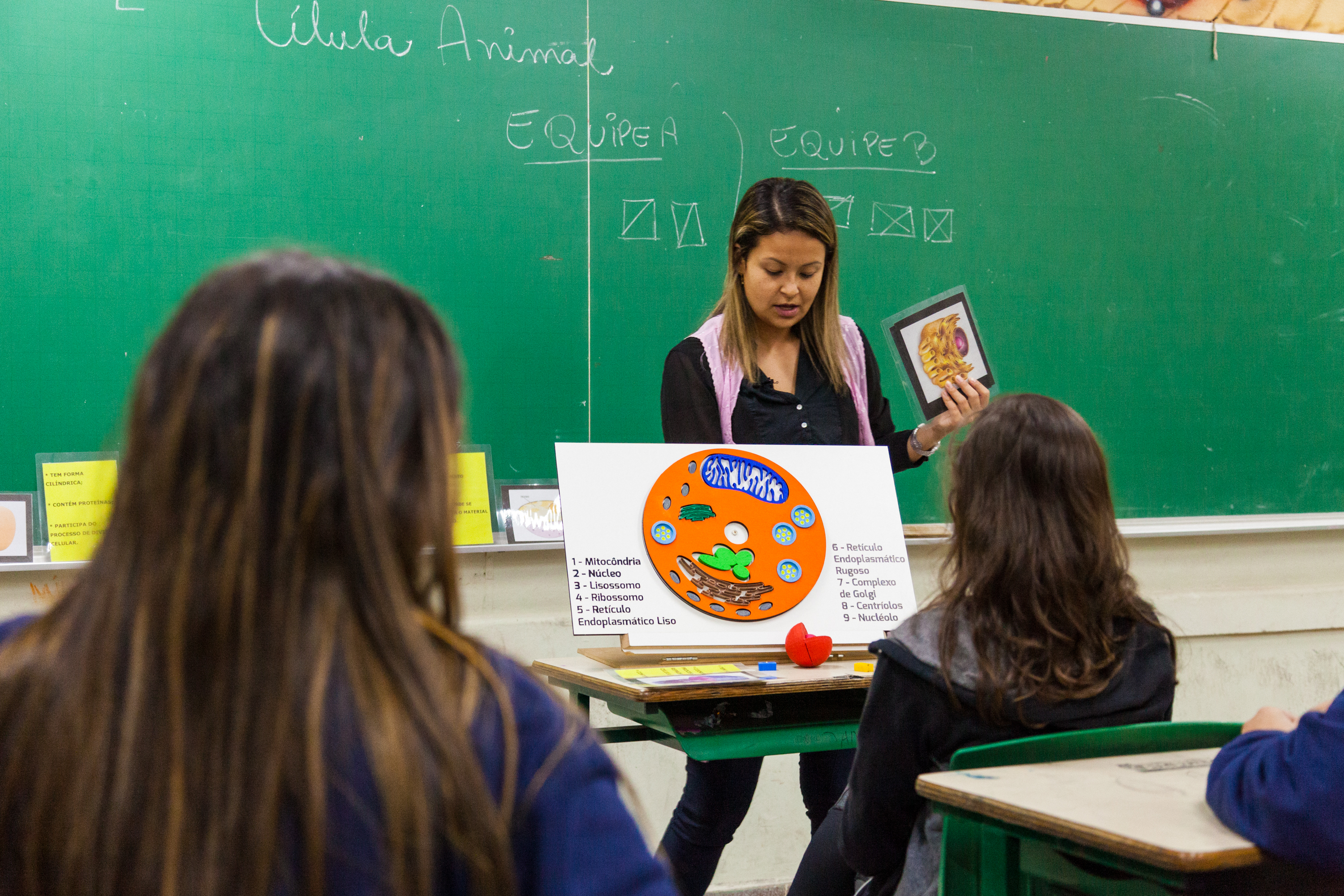 Em sala de aula e em frente à lousa, professora apresenta material pedagógico acessível Célula Tátil para a turma. Fim da descrição.
