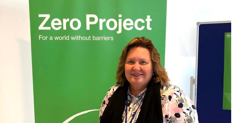 Deborah Dunstone posa para foto em frente a cartaz em que se lê: "Zero Project: for a Deborah Dunstone posa para foto em frente a cartaz em que se lê: "Zero Project: for a world without barriers". Fim da descrição.