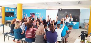 Em sala de aula, educadores e familiares de estudantes sentam em cadeiras e levantam os braços em votação. Fim da descrição