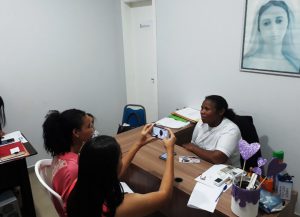 Em escritório, duas estudantes entrevistam Maria das Dores Pereira Anjos. Uma delas segura um celular, gravando o rosto de Maria das Dores. Na parede, está pendurado um quadro de Nossa Senhora Aparecida. Fim da descrição.