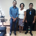 De pé, a juíza Tania Vasconcellos posa para foto sorridente com dois estudantes em seu escritório. Fim da descrição.