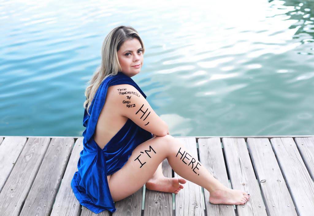 Sentada em deck de madeira, Tathi posa foto com um vestido azul. Em seu braço e perna direta está escrito: "Hi, I'm here" e "Tathi Piancastelli by Gabi Artz". Fim da descrição. 