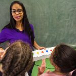 Em sala de aula, professora conversa com duas alunas, enquanto segura a caixa da lousa interativa em sua mão esquerda. Fim da descrição.
