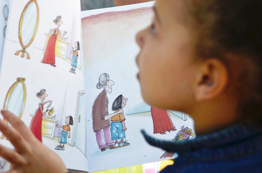 A foto foca um livro com ilustrações de uma mulher e de uma menina. Uma criança, que aparece desfocada, segura o livro e olha para cima. Fim da descrição.
