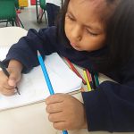 Em sala de aula, e sentada em cadeira, Jhacel pinta em caderno apoiado em mesa escolar. Fim da descrição.