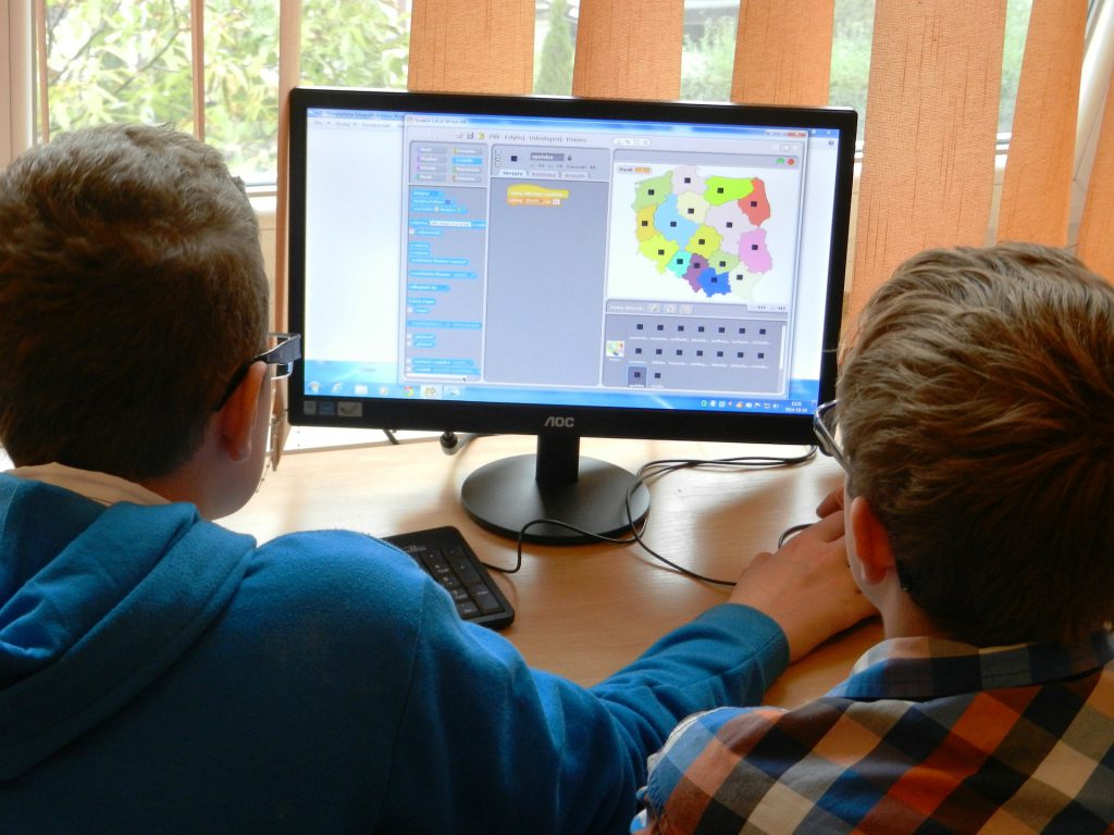 Duas crianças usando óculos estão em frente a computador que mostra um mapa colorido por região. Uma delas está segurando o mouse. Fim da descrição.