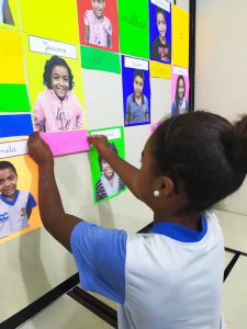 Em painel colorido com fotos de vários estudantes exposto em sala de aula, aluna coloca elogio escrito em papel rosa na foto de colega de classe. Fim da descrição.