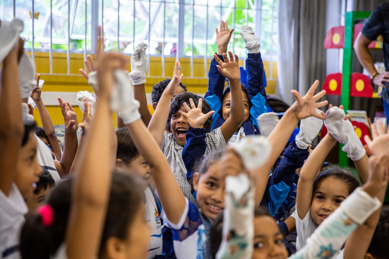 Em sala de aula, estudantes levantam as mãos e sorriem. Todos usam as luvas da Cartola mágica em uma das mãos. Fim da descrição.