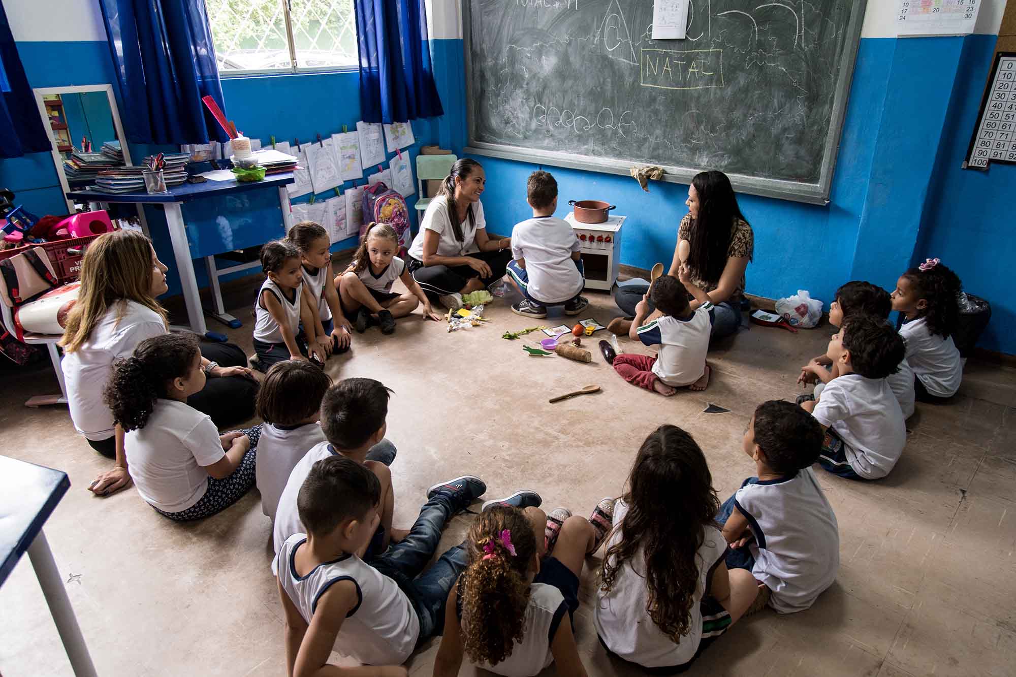 Em sala de aula, estudantes em roda sentados no chão observam aluno manuseando fogão musical, enquanto duas professoras supervisionam a atividade. Fim da descrição.