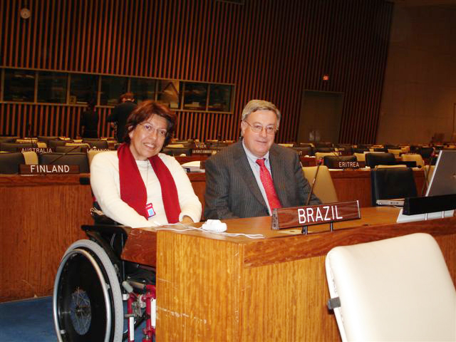 Em plenário da ONU, Izabel Maior, em cadeira de rodas, e Don MacKay, presidente do Comitê de Elaboração da Convenção, posam para foto atrás de mesa do plenário, sobre a qual uma placa em que se lê "Brazil". Fim da descrição
