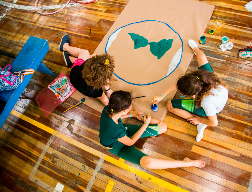 Sentadas em chão de madeira, três meninas usam pincel e tinta guache para pintar papel pardo. É possível identificar o início de um mapa mundi, estando já desenhado um círculo em azul, o formato do continente americano em verde e os pólos sul e norte em branco. Fim da descrição.
