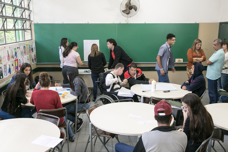 Em sala de aula, estudantes se organizam ao redor de mesas circulares. Em frente à lousa, educadores conversam com estudante. Fim da descrição.