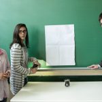 Em frente a papel colocado em lousa, três estudantes observam sala de aula. Fim da descrição.