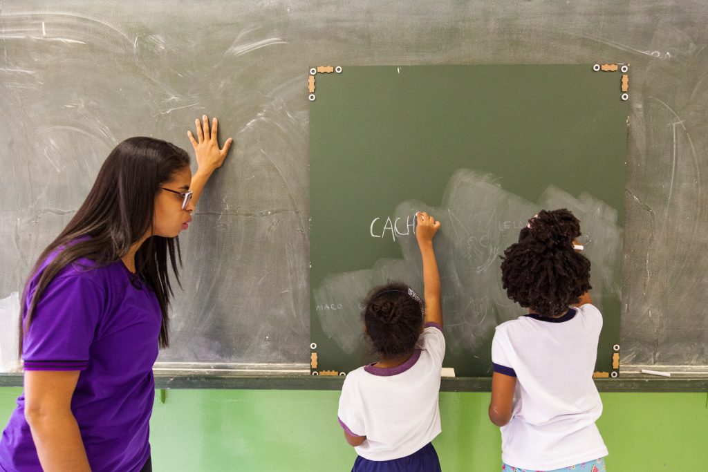 Duas alunas escrevem com giz branco na lousa interativa, que está apoiada no quadro negro da sala de aula, enquanto professora as observa. Fim da descrição.
