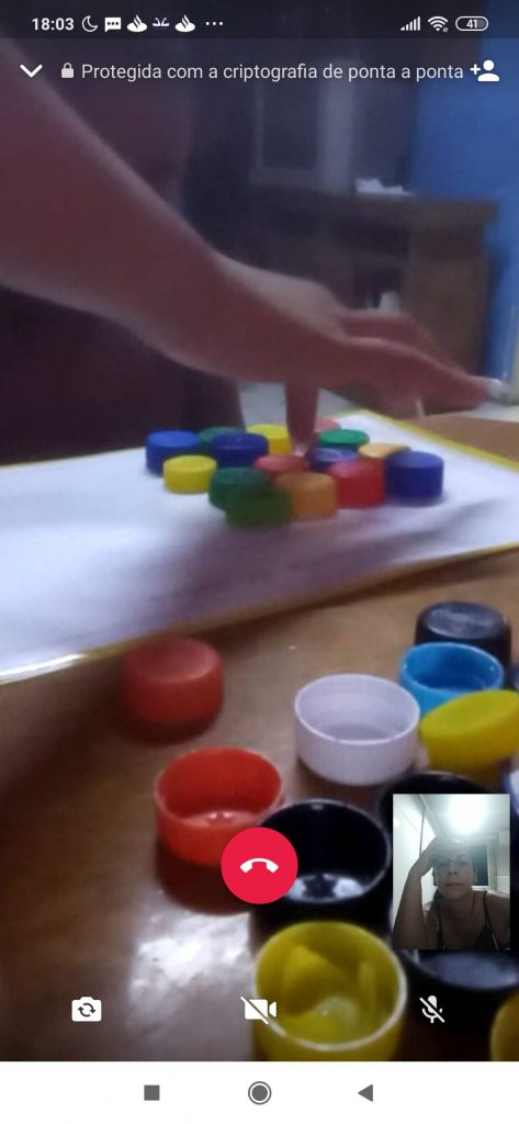 Em videochamada, professora Vilma, em quadrado menor no canto inferior direito da tela, observa o estudante realizar uma atividade com tampinhas coloridas. Fim da descrição.