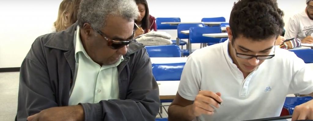 Em sala de aula, um estudante na faixa dos 60 anos e com óculos escuros observa atento estudante escrevendo ao seu lado. Fim da descrição.